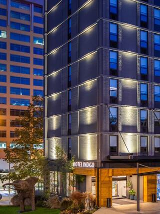Exterior view of Hotel Indigo Chattanooga at dusk, featuring a modern gray-brick facade with accent lighting, large blue-tinted windows, and a warmly lit entrance framed by wood paneling, with sculptural landscaping in the foreground.