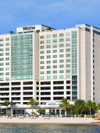 Waterfront exterior of The Westin Tampa Bay, a modern high-rise hotel featuring glass windows, private balconies, palm trees, and AQUA restaurant, overlooking the white sand beach and bay views.