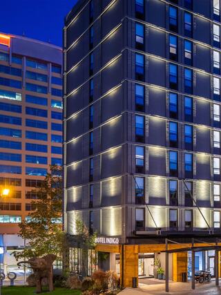 Exterior view of Hotel Indigo Chattanooga at dusk, featuring a modern gray-brick facade with accent lighting, large blue-tinted windows, and a warmly lit entrance framed by wood paneling, with sculptural landscaping in the foreground.