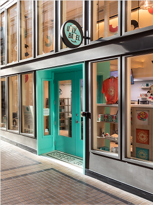 Exterior storefront of Ugly Bagel featuring a vibrant turquoise entrance, modern retail display windows, and contemporary branding in a stylish urban setting.