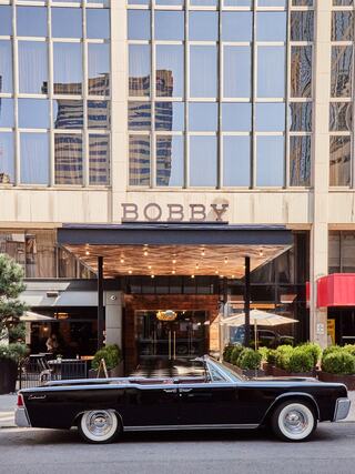 Front entrance of Bobby Hotel in downtown Nashville featuring a vintage black Lincoln Continental parked curbside, with large modern windows reflecting the city skyline above the marquee.