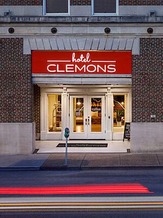 Street view of the entrance to Hotel Clemons in Chattanooga, featuring a red sign with white lettering above double glass doors, flanked by large windows and set in a brick building facade.